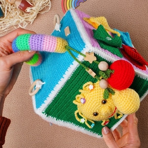 May include: A colourful crocheted cube toy with different textures and activities for babies and toddlers. The cube features a yellow crocheted character with a smiling face, a red crocheted ball, and a rainbow-coloured crocheted loop.