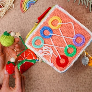 May include: A colorful crocheted activity cube with various textures and shapes for babies and toddlers. It features a variety of loops, a watermelon, a lime, a strawberry, and a small crocheted animal.