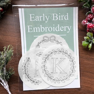 May include: An embroidery kit in a clear plastic sleeve. The kit includes circular designs with letters and floral wreaths. The text "Early Bird Embroidery" is visible. The background features pine and red berries.
