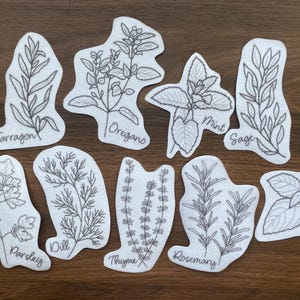 Op de afbeelding: Negen witte viltkruidenuitsparingen met zwarte lijntekeningen van kruiden, waaronder dragon, oregano, munt, salie, peterselie, dille, tijm, rozemarijn en basilicum. Elke uitsparing heeft de naam van het kruid in handschrift.