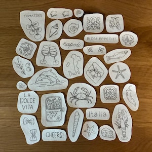 May include: A collection of small, irregularly shaped patches with hand-drawn illustrations and text. Designs include tomatoes, wine glasses, a crab, a fish, and the words "Bon Appetit!" and "Italia."