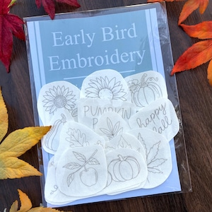 Könnte beinhalten: Eine durchsichtige Kunststoffverpackung mit mehreren weißen Stoffaufnähern, die schwarze Linienstickereien von Sonnenblumen, Kürbissen, Blättern sowie den Text "Pumpkin Patch" und "happy fall" auf einer blauen Karte mit der Aufschrift Early Bird Embroidery zeigen.
