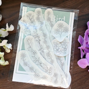 Könnte beinhalten: Eine Packung weißer Nagelkunst-Aufkleber mit floralen Designs. Die Aufkleber in verschiedenen Formen haben zarte schwarze Strichzeichnungen von Blumen und Blättern. Die Verpackung ist in klarem Kunststoff versiegelt, mit einem hellgrünen und weißen Hintergrund. Weiße und lila Blumen umgeben die Packung.