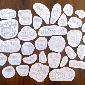 May include: A collection of white fabric patches with gray line drawings of autumn-themed images, including pumpkins, apples, leaves, and the word "autumn". Other patches say "Pumpkin Season", "Autumn Vibes", "Autumn is Here", and "Cozy Season".