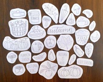 May include: A collection of white fabric patches with gray line drawings of autumn-themed images, including pumpkins, apples, leaves, and the word "autumn". Other patches say "Pumpkin Season", "Autumn Vibes", "Autumn is Here", and "Cozy Season".