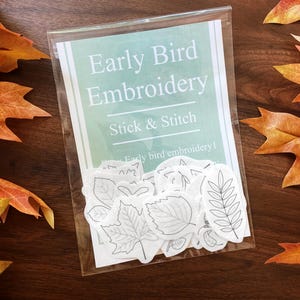 May include: A clear plastic package containing "Early Bird Embroidery" stick and stitch leaf designs. The package is surrounded by autumn leaves in shades of orange, red, and yellow, set on a dark wooden surface.
