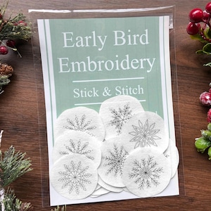 May include: A package of "Early Bird Embroidery Stick & Stitch" featuring several white, circular patches with black snowflake designs. The package is clear, and the background is a wooden surface with decorative greenery.