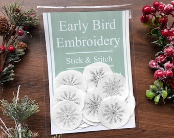 May include: A package of "Early Bird Embroidery Stick & Stitch" featuring several white, circular patches with black snowflake designs. The package is clear, and the background is a wooden surface with decorative greenery.
