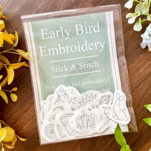 May include: A clear plastic package containing an "Early Bird Embroidery Stick & Stitch" kit. The kit includes a sheet with various small, black and white embroidery designs, including a whale, sun, and bicycle. The background is a wooden surface with yellow and green foliage.