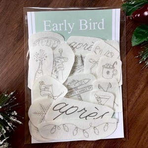 May include: A collection of off-white fabric patches featuring black line art illustrations of winter and holiday themes, including a champagne bottle, a mug, a snowflake, and the text "Early Bird" and "après" inside a clear plastic sleeve.
