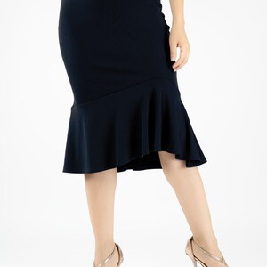 May include: A black midi skirt with a ruffled hem. The skirt is fitted through the hips and thighs and flares out at the bottom. The model is wearing the skirt with a pair of gold heels.