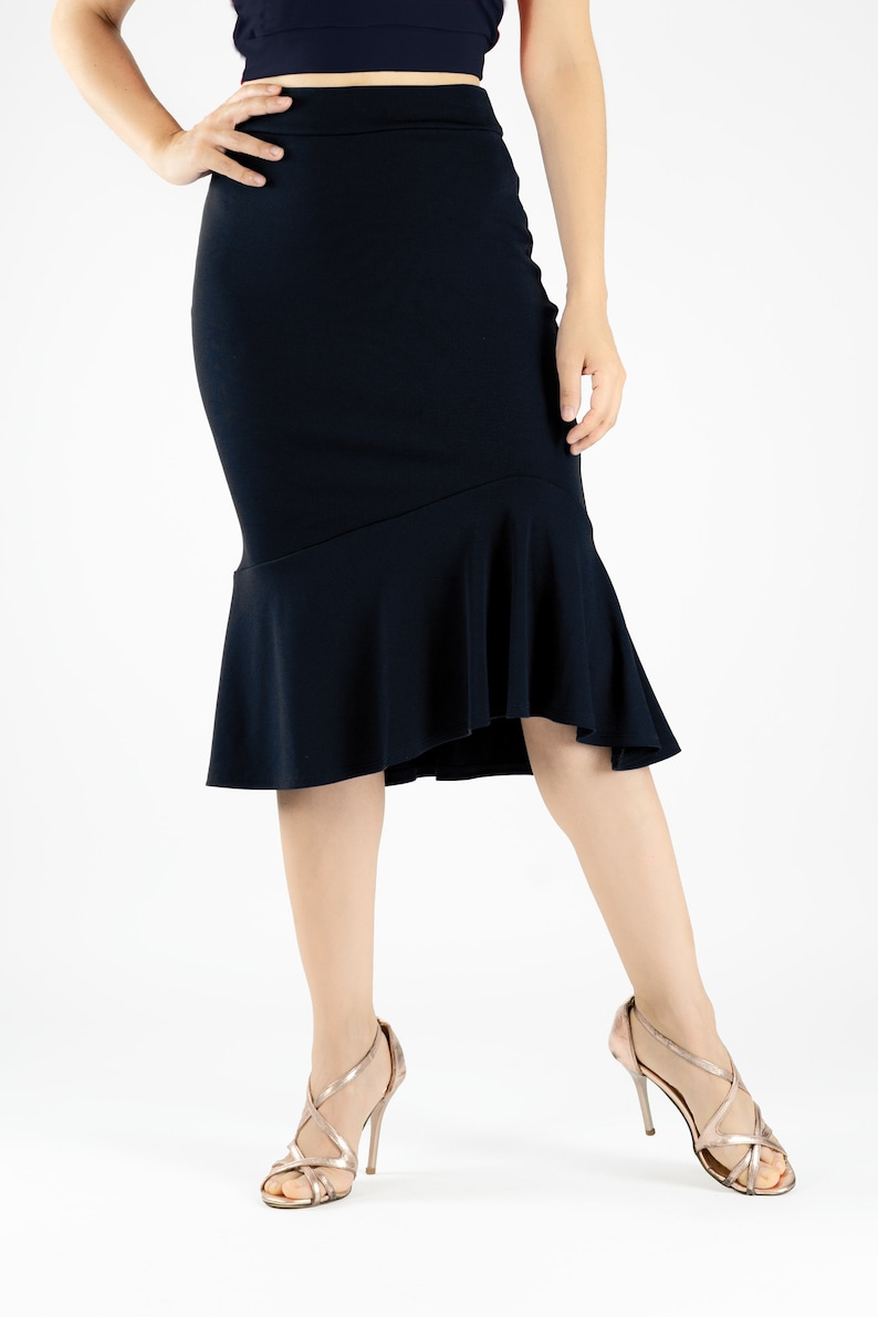 May include: A black midi skirt with a ruffled hem. The skirt is fitted through the hips and thighs and flares out at the bottom. The model is wearing the skirt with a pair of gold heels.