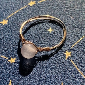 May include: A delicate gold wire ring with a white gemstone. The ring is on a blue background with gold stars.