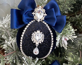 May include: A navy blue Christmas ornament with a velvet texture, adorned with a silver-colored jewel and a matching bow. The ornament is embellished with a silver-colored trim and a teardrop-shaped jewel. The background features frosted pine branches.