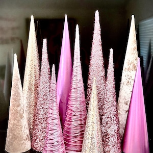 Könnte beinhalten: Eine Sammlung von rosa und weißen, kegelförmigen Weihnachtsbaumschmuck. Die Bäume sind aus verschiedenen Materialien gefertigt, darunter Samt, Pailletten und Stoff.