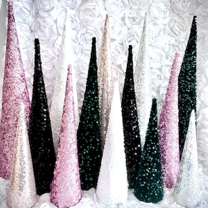 May include: Assortment of sequined cone-shaped Christmas trees in various colors, including pink, white, black, and green. The trees are arranged in a row on a white background.