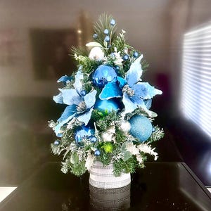 Puede incluir: Centro de mesa navideño festivo con un pequeño arreglo de ramas de pino artificiales, flores de Pascua azules y adornos plateados y azules. El arreglo está colocado en un recipiente decorativo plateado y blanco, creando una exhibición navideña.