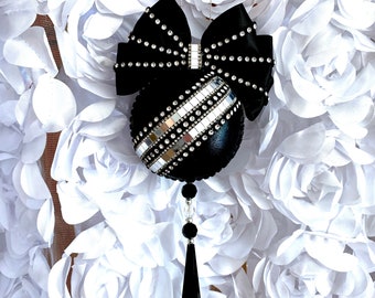 May include: Black Christmas ornament with a silver and black striped design and a black bow with rhinestones. The ornament is hanging from a white background.