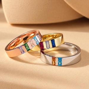 May include: Three metal rings in rose gold, yellow gold, and silver tones. Each ring features a flat band design with embedded rectangular baguette-cut gemstones in various colors including blue, green, pink, purple, red, and orange.