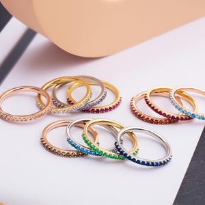 May include: A set of thin gold rings with colorful gemstones. The rings are stacked and arranged on a white surface.