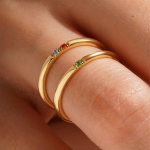 Könnte beinhalten: Zwei goldene Ringe mit kleinen bunten Edelsteinen. Der obere Ring hat einen roten, blauen und grünen Edelstein. Der untere Ring hat einen grünen Edelstein.