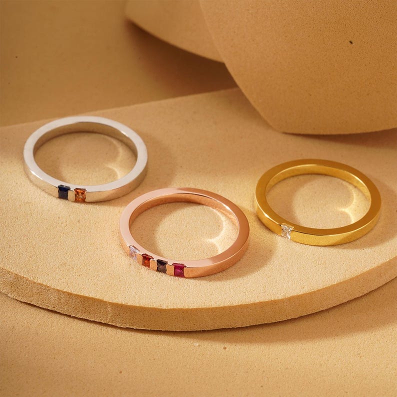 Pode incluir: Tr&ecirc;s bandas simples de ouro, ouro rosa e prata com pedras preciosas coloridas. Cada banda tem uma fileira de pequenas pedras preciosas quadradas.