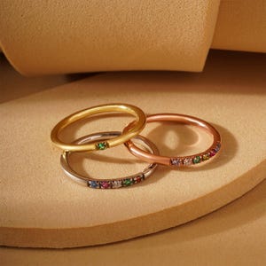 Peut inclure: Trois fines bagues en or avec des pierres précieuses colorées. Les bagues sont empilées les unes sur les autres. La bague du haut est en or jaune avec une seule pierre précieuse verte. La bague du milieu est en or rose avec une rangée de pierres précieuses colorées. La bague du bas est en or blanc avec une rangée de pierres précieuses colorées.