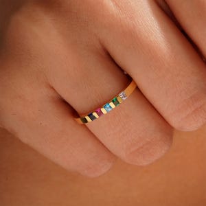 Pode incluir: Um anel de banda de ouro com uma fileira de pedras quadradas coloridas e uma &uacute;nica pedra quadrada transparente no centro.