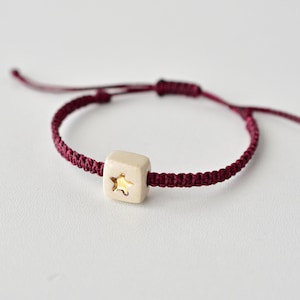 Puede incluir: Un brazalete de cuerda de color rojo burdeos con una cuenta cuadrada blanca con una estrella dorada.