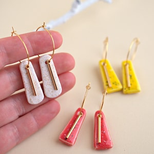 Può includere: Orecchini a cerchio dorati con pendenti rettangolari bianchi strutturati e barre dorate. Sono visibili anche altri orecchini gialli e rossi con barre dorate. Gli orecchini hanno un design moderno e minimalista.