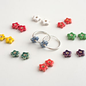 May include: A pair of silver hoop earrings with blue star-shaped ceramic charms. The charms have a white speckled glaze and are surrounded by other colorful star-shaped charms in red, yellow, green, and purple.