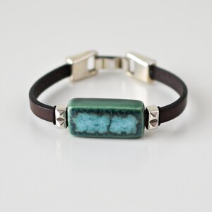 Op de afbeelding: Een bruin leren armband met een zilveren sluiting en een rechthoekige groene en blauwe keramische kraal.