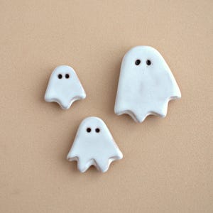 Op de afbeelding: Drie witte keramische knopen in de vorm van een geest met gladde, glanzende oppervlakken. Elke geest heeft twee kleine, donkere, ronde inkepingen als ogen. De set bevat één grotere geest en twee kleinere geesten met golvende onderranden.