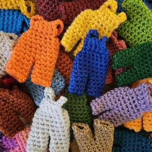 May include: A collection of colorful, miniature crochet overalls in various shades, including orange, yellow, blue, green, and purple. The handmade items are arranged in a close-up shot, showcasing the texture and detail of the yarn.