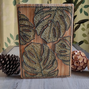 Peut inclure: Décoration murale en bois représentant des feuilles de Monstera en fil tendu. Les feuilles sont réalisées avec du fil doré et vert sur un fond en bois brun clair. L'œuvre est encadrée d'une bordure marron foncé, créant un thème tropical.