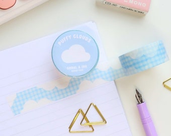 May include: A desk scene featuring stationery items. A roll of "Puffy Clouds" washi tape with a blue and white cloud pattern is unrolled on a lined notepad. Other items include a pink eraser, a purple pen, gold paper clips, and a pink keyboard.