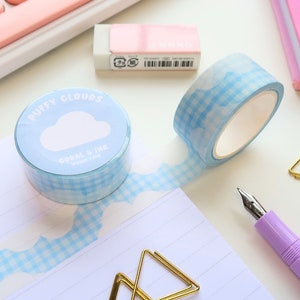 May include: A roll of light blue washi tape with a cloud design and the text "Puffy Clouds". The tape has a gingham pattern and is unrolled on a white surface. A pink eraser, a pink notebook, and a gold paperclip are also visible.