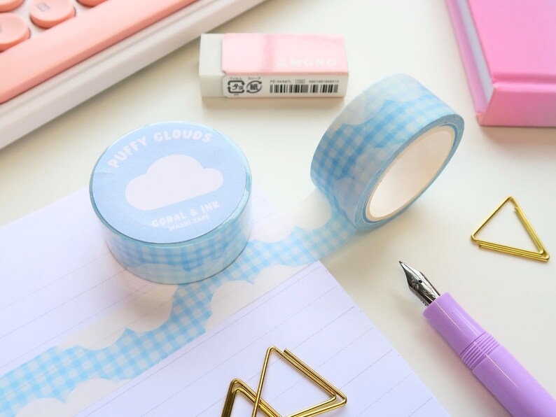 May include: A roll of light blue washi tape with a cloud design and the text "Puffy Clouds". The tape has a gingham pattern and is unrolled on a white surface. A pink eraser, a pink notebook, and a gold paperclip are also visible.