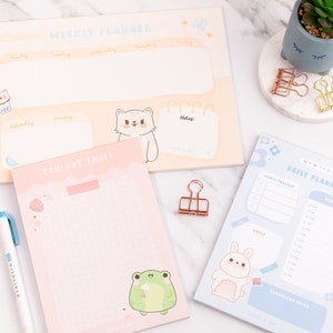May include: Three different pastel coloured notepad planners with cute animal illustrations. The weekly planner is yellow with a cat illustration, the daily planner is blue with a bunny illustration, and the to-do list notepad is pink with a frog illustration. The text on the planners reads "Weekly Planner", "Daily Planner", and "You Got This!"