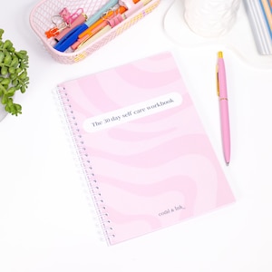 Może przedstawiać: R&oacute;żowy zeszyt w spirali z ćwiczeniami dotyczącymi samoopieki z tytułem "The 30 day self care workbook" i tekstem "coral & ink" na okładce.