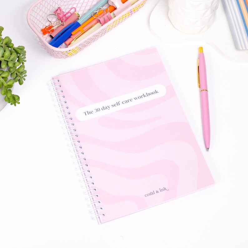 Może przedstawiać: R&oacute;żowy zeszyt w spirali z ćwiczeniami dotyczącymi samoopieki z tytułem "The 30 day self care workbook" i tekstem "coral & ink" na okładce.