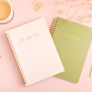 May include: Two spiral bound notebooks on a pink surface. One notebook is pink with the words "Milk Tea" written in gold lettering. The other notebook is green with the words "Matcha" written in gold lettering.