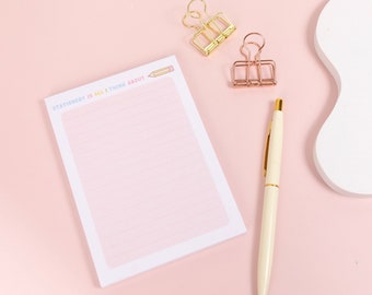 May include: A pink notepad with lined pages and the text "STATIONERY IS ALL I THINK ABOUT" is next to a gold pen and two binder clips. The notepad has a pencil graphic. The background is a light pink.