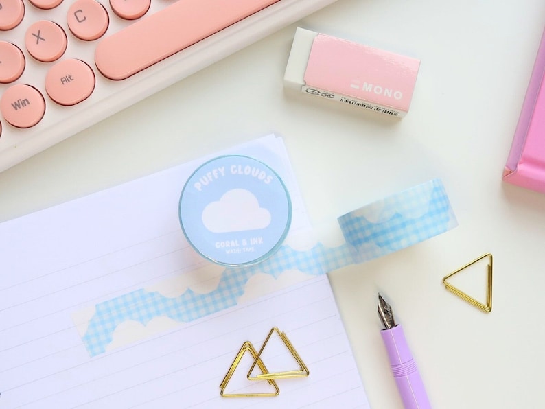 May include: A desk scene with stationery. A roll of "Puffy Clouds" washi tape with a blue and white cloud pattern is unrolled on a lined notepad. Other items include a pink eraser, a purple pen, gold paper clips, and a pink keyboard.