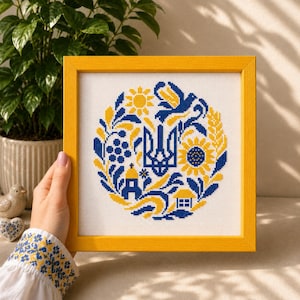 May include: A square yellow frame containing a cross-stitch embroidery on white fabric. The design features a blue and yellow circular pattern with a central trident symbol, a sunflower, a bird, a small house, and a church, all in blue and yellow.