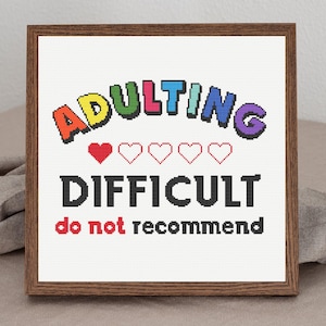 May include: A framed cross-stitch design with the text "Adulting Difficult do not recommend". The text "Adulting" is in rainbow colors and the text "Difficult" is in black. There are five red hearts below the word "Adulting".