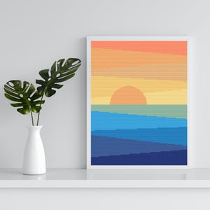 May include: A framed cross-stitch artwork depicts a sunset over the ocean. The sky transitions from orange to yellow, with a round sun. The ocean is rendered in shades of blue. A white vase with green leaves is on the left.