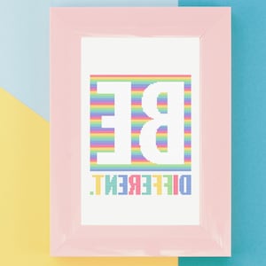 May include: A framed cross-stitch artwork with a pink frame. The artwork features the word "BE" in large white letters above the word "DIFFERENT" in green and yellow letters. The letters are set against a rainbow striped background.