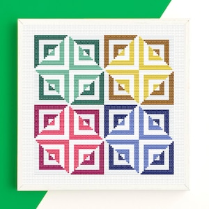 Puede incluir: Una obra de arte de punto de cruz enmarcada con un diseño geométrico. La obra presenta nueve cuadrados, cada uno con un patrón en forma de cruz de cuadrados anidados en varios colores, incluyendo verde, rosa, azul y amarillo. Fondo blanco.