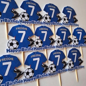 May include: Blue soccer jersey cupcake toppers with the name "FREDDIE" and the number "7". Each topper includes a soccer ball and a pair of black cleats. The words "Happy Birthday" are printed on a banner.